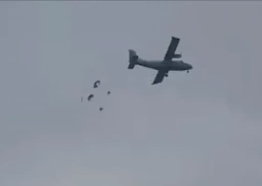 A turboprop aircraft flying against a grey sky with several objects descending beneath it, each attached to small parachutes.